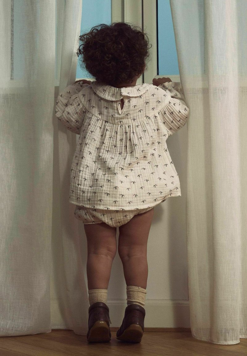 Šviesios spalvos, tekstūruoto audinio marškiniai su apvaliu apykakle ir mažais raštais. Dera su atitinkančiomis kelnaitėmis ir tamsiais batais. Vaikas stovi šalia lango.