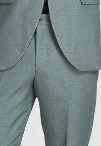 Completo grigio chiaro con giacca su misura e pantaloni a gamba dritta. Il tessuto ha una consistenza liscia e una leggera lucentezza, con tagli classici.