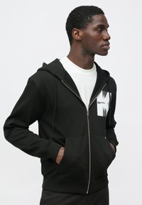 Μαύρο φούτερ με φερμουάρ, κατασκευασμένο από μαλακό ύφασμα, με μπροστινή τσέπη, ριμπ μανσέτες και λευκό γραφικό σχέδιο στην αριστερή πλευρά του στήθους.