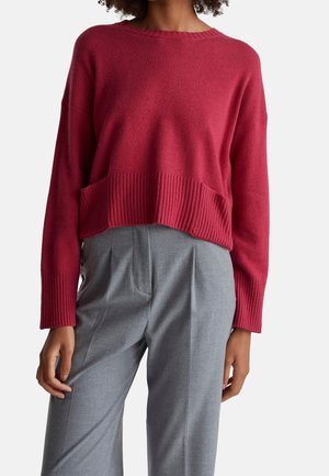 Femme portant un pull rouge ample à manches longues avec ourlet et poignets côtelés, associé à un pantalon plissé gris taille haute.