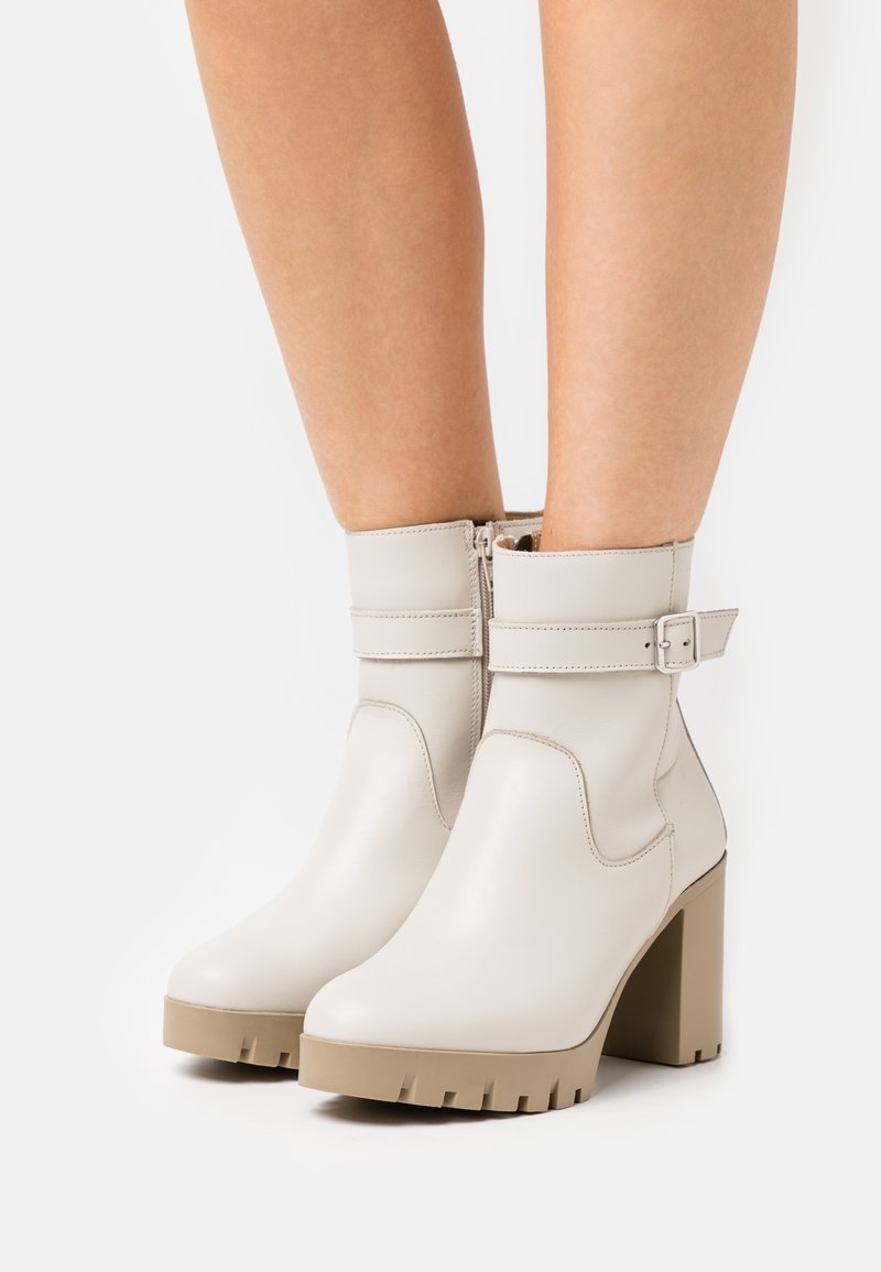 Stivaletti alla caviglia color crema realizzati in pelle liscia, con un tacco a piattaforma beige spesso e una cintura regolabile con una fibbia argentata.