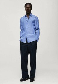 Camicia button-up a righe blu chiaro realizzata in cotone, abbinata a pantaloni scuri su misura. La camicia ha un colletto classico e una silhouette aderente.