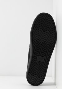Svart sneaker med en texturerad gummisula som har ett vågmönster, slätt yttermaterial och en rektangulär logotyp med texten "TOMS."