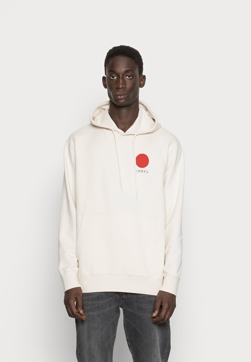 Felpa color crema con tasca frontale, cappuccio con lacci e logo circolare rosso sul lato sinistro del petto, indossata con jeans grigi.