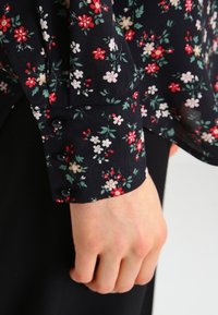 Blusa nera a fiori con un motivo di fiori rossi, bianchi e verdi; presenta un polsino con bottoni e una texture di tessuto morbido e leggero.