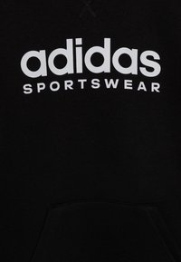 Musta collegepaita kengurutaskulla. Valkoinen "adidas SPORTSWEAR" -logo näkyvästi rinnassa, pehmeästä puuvillamateriaalista valmistettu.