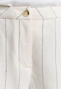 Witte stoffen broek met zwarte verticale strepen, voorzien van een sluiting met een voorste knoop en een subtiel zakdetail aan de linkerkant.