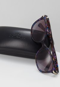 Ochelari de soare tortoise violet cu lentile negre degradate, accesorii aurii pe brațe și un etui Coach negru cu logo embosat.