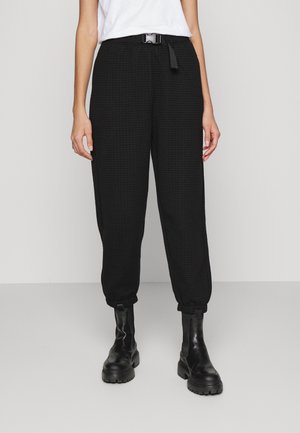 Henkilö, jolla on päällään teksturoidut mustat housut vyöllä, valkoinen takki sisällä ja mustat paksupohjaiset nahkanilkkurit, seisomassa harmaalla lattialla.