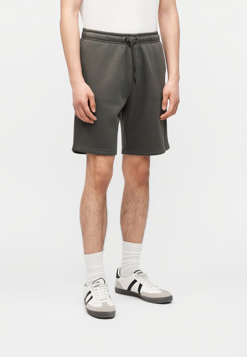 Homme portant un short gris foncé à cordon, des chaussettes blanches montantes et des baskets blanches avec des rayures noires, debout devant un fond uni.