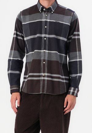 Mężczyzna w długim rękawie, zapinanej na guziki koszuli z dużym szaro-czarnym i brązowym wzorem w kratę oraz ciemnobrązowych spodniach ze sztruksu.
