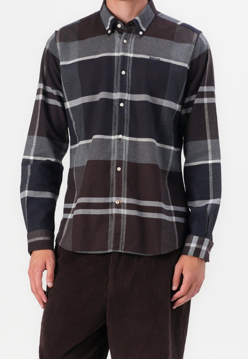 Homme portant une chemise boutonnée à manches longues avec un grand motif à carreaux gris, noir et marron et un pantalon en velours côtelé marron foncé.
