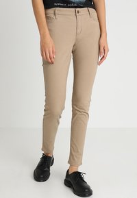 Slim-fit beige byxor bärs med svarta oxfordskor och en svart topp med delvis vit text på en ljus bakgrund.