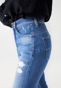 Modré džíny z denimu mají přiléhavý střih s vysokým pasem, opotřebované detaily na kolenou a decentní šití.