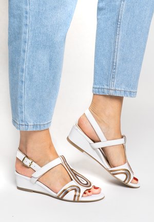 Füße in weißen Keilsandalen mit goldenen und braunen Streifen, kombiniert mit hellblauen Jeans, die oberhalb der Knöchel gekürzt sind.