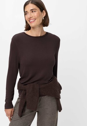 Lächelnde Frau, die ein dunkelbraunes Langarmshirt trägt, mit einem Strickpullover um die Taille gebunden und karierte Hose.