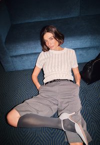 Gai beidzas adīts tops ar svītrotu rakstu, pelēkas svītrainas platas bikses, apvienotas ar pelēkām un baltām augstpapēžu kurpēm uz teksturēta zila paklāja.