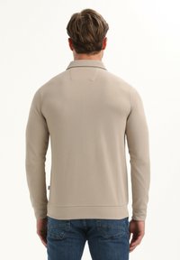 Beżowy sweter z długimi rękawami i kołnierzykiem, o gładkiej fakturze i prążkowanych mankietach. W połączeniu z ciemnoniebieskimi dżinsami.