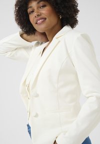 Blazer blanc ajusté avec revers crantés, fermetures à boutons et poches latérales. Tissu lisse avec une silhouette cintrée et un design moderne.
