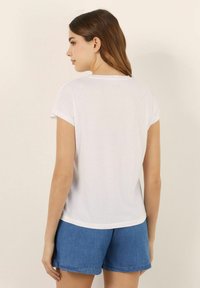 Tricou alb cu mânecă scurtă și manșete răsucite, textură moale, decolteu rotund și croială ușor largă. Asortat cu pantaloni scurți din denim.