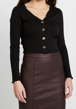 Crop top noir côtelé avec un décolleté en V et cinq boutons ronds marron sur le devant. Associé à une jupe en cuir marron taille haute.