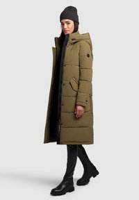 Olive green puffer coat with a hood, zip and button closure, side pockets, and black accents; paired with black leggings and ankle boots.