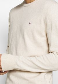 Beige lehký svetr vyrobený z měkké tkaniny, s kulatým výstřihem, žebrovanými manžetami a malým logem na hrudi.