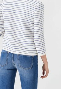 Pitkähihainen, valkoinen ja sininen raitapaita; teksturoitu kangas. Yhdistetty sinisiin farkkuihin, joissa näkyvät taskut ja hienovaraiset ompelu- ja yksityiskohdat.