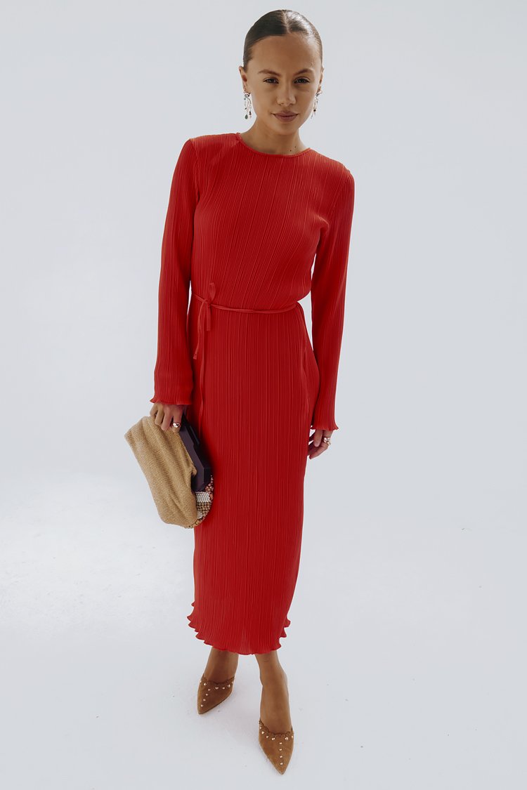 Abito maxi rosso plissettato con maniche lunghe, vita cintrata e orlo arricciato. Abbinato a una pochette beige e décolleté nude con punte e borchie.
