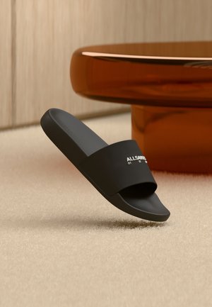 Sandale à glissière noire de marque sur un tapis texturé beige avec une table ronde ambrée-brune en arrière-plan.