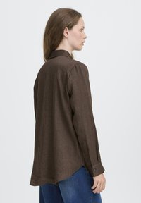 Eine Frau im Profil, die ein lockeres braunes langärmliges Shirt und blaue Jeans trägt, steht vor einem schlichten hellen Hintergrund.