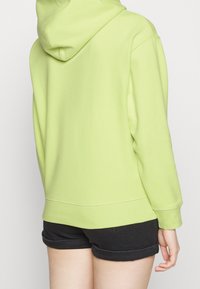 Sudadera con capucha de color verde lima, hecha de una tela suave, con un bolsillo tipo canguro en la parte frontal y puños acanalados, combinada con pantalones cortos negros.