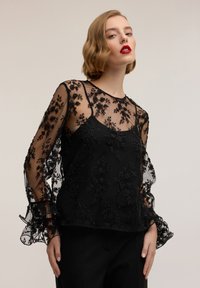 Blusa nera con motivi floreali ricamati trasparenti e dettagli con perline. Presenta maniche lunghe e fluide e una scollatura rotonda.