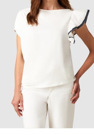 Kobieta ubrana w biały top z krótkim rękawem, ozdobiony falbanką i ciemnym wykończeniem na jednym ramieniu, w połączeniu z białymi spodniami, ręce spoczywają po bokach.