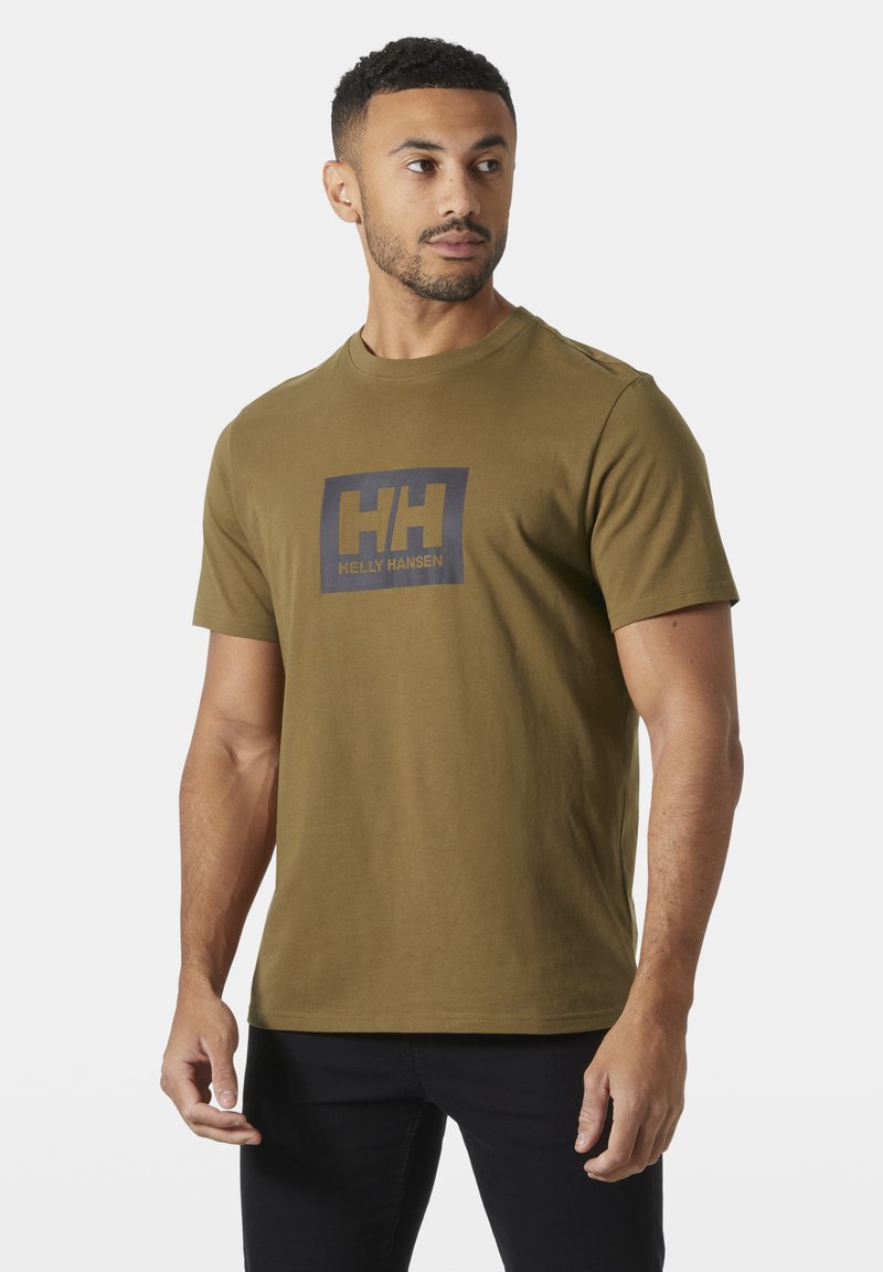 Hombre con camiseta Helly Hansen verde oliva y pantalones negros, mirando hacia su izquierda contra un fondo claro y liso.