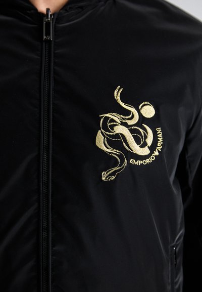 Casaco bomber preto de nylon com fecho ao centro, apresentando um logotipo dourado bordado em destaque e detalhes de design no peito.