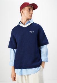 Jeune homme portant un pull court à manches courtes bleu marine avec le texte "Originals Club" sur une chemise à rayures bleu clair et une casquette rouge, les mains dans les poches.
