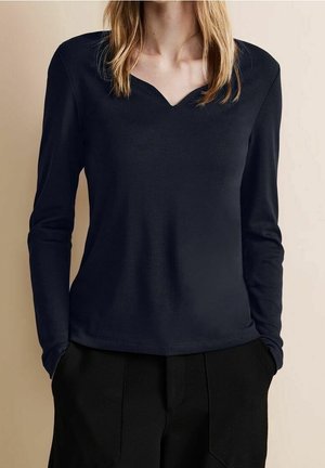 Nainen pukeutuneena pitkähihaiseen mustaan toppiin, jossa on lovellinen pääntie, ja mustiin korkeavyötäröisiin housuihin, kädet taskuissa, beige tausta.