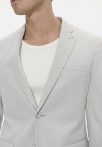 Light gray blazer made from textured fabric, featuring a single button closure and a chest pocket, worn over a white ribbed shirt.