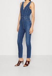 Mono de mezclilla con escote en V profundo, diseño ajustado y piernas acampanadas. Presenta costuras frontales y un acento plateado en el cuello.