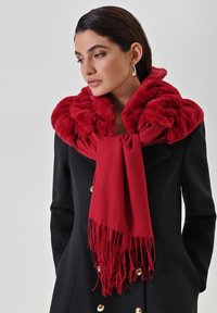 Sciarpa in pelliccia sintetica rossa con un design arricciato, rifinita con bordi frangiati, abbinata a un cappotto nero con bottoni dorati.