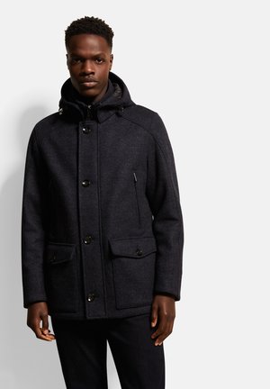 Dunkler Wollmantel mit Kapuze, mit einer Knopfleiste vorne, zwei großen Fronttaschen und einer strukturierten Oberfläche. Akzente sind schwarze Knöpfe.
