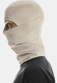 Person wearing a beige balaclava covering head and lower face, with a black shirt, shown in profile against a plain background.
