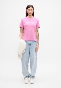 Rozā kokvilnas t-krekls ar "Calvin Klein Jeans" logo, saskaņots ar gaiši ziliem oversized džinsiem un baltiem apaviem, turot rokās smilšu krāsas džemperi.