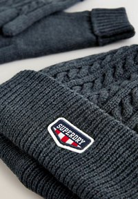 Graue gestrickte Mütze mit einem strukturierten Kabelmuster und einem gerippten Saum, versehen mit einem Patch mit der Aufschrift "Superdry" in Blau, Weiß und Rot.