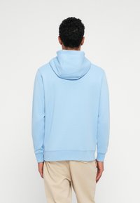 Homme aux cheveux bouclés portant un sweat à capuche bleu clair et un pantalon beige, debout de dos face à la caméra sur un fond blanc uni.
