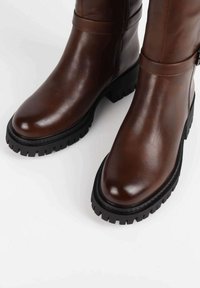 Bottes hautes jusqu'au genou en cuir marron à finition lisse, bout arrondi, semelle en caoutchouc noire épaisse, avec une bride autour de la cheville.