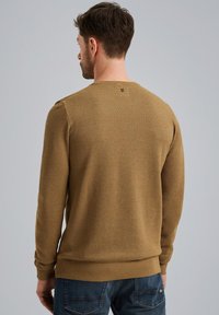 Brązowy sweter o fakturze z okrągłym dekoltem i prążkowanymi mankietami, zestawiony z niebieskimi dżinsami. Wyjątkowy detal przeszycia na plecach.