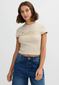 Camiseta corta de canalé beige con mangas cortas, que presenta un diseño de texto en marrón claro que dice "Sé una voz, no un eco". Combinada con jeans azules.