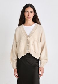 Beige cropped cardigan with V-neckline, ribbed edges, and two front pockets. Features three large buttons and soft knit texture.